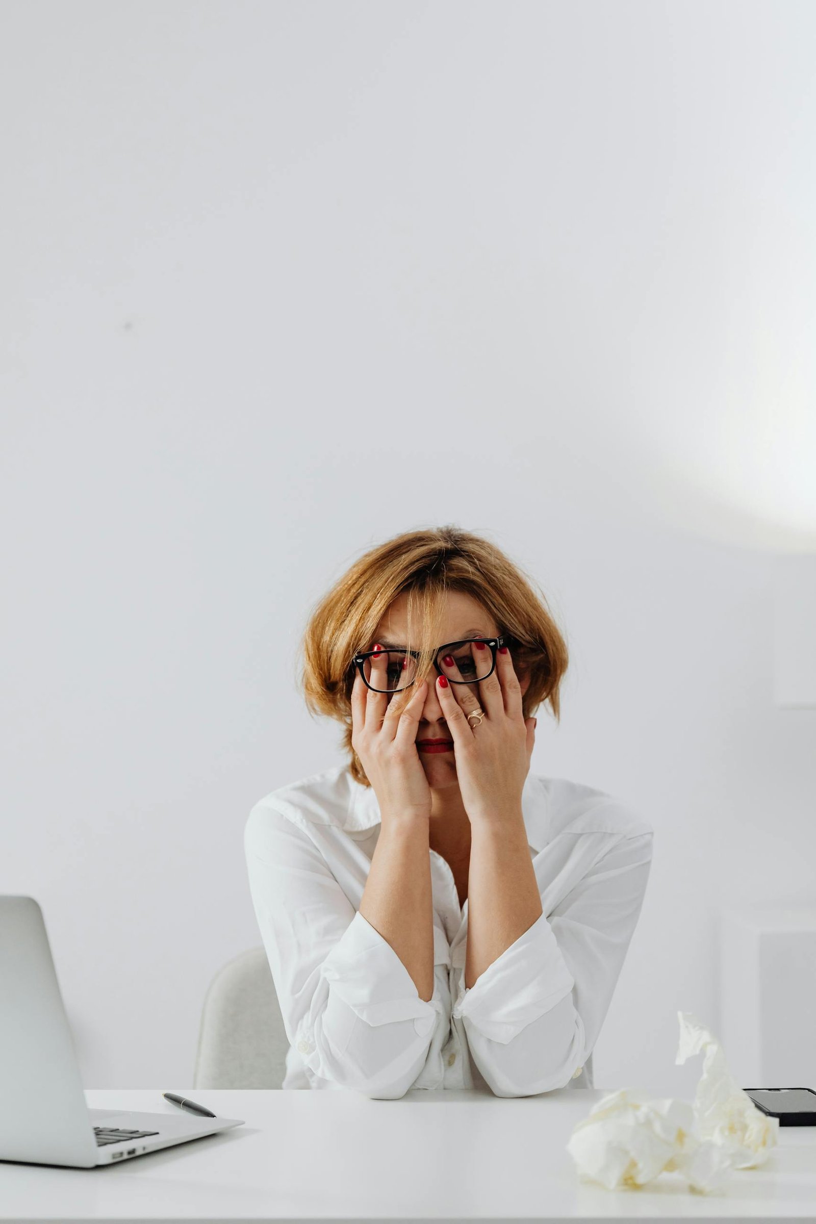 A stressed woman in eyeglasses and white shirt sits at her desk, overwhelmed and exhausted.